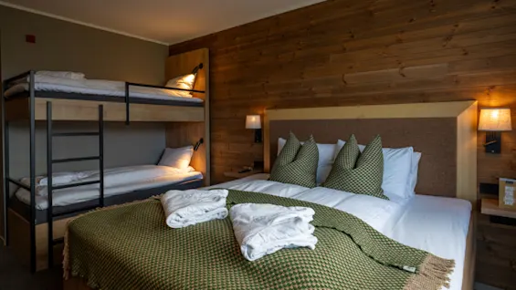 Ett mysigt hotellrum med en stor säng med gröna plädar och kuddar, samt en våningssäng i bakgrunden.