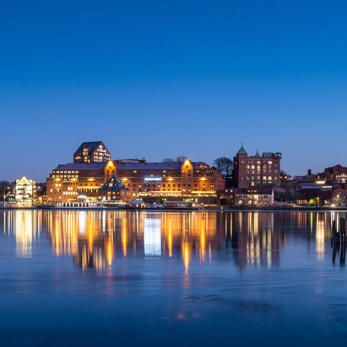Buildings brightly illuminated at night reflect in calm water, with a clear sky above. A large waterfront building has a sign: "Clarion Hotel Wisby."