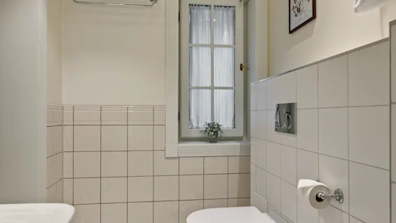 Et lyst og rent badeværelse med toilet, vindue med gardiner og flisebelagte vægge. Et billede mærket FLOWERS hænger på væggen.