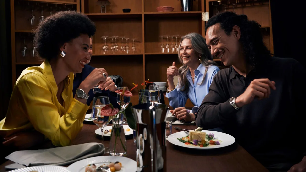 Four people dining and laughing together at a restaurant table elegantly set with food and drinks.