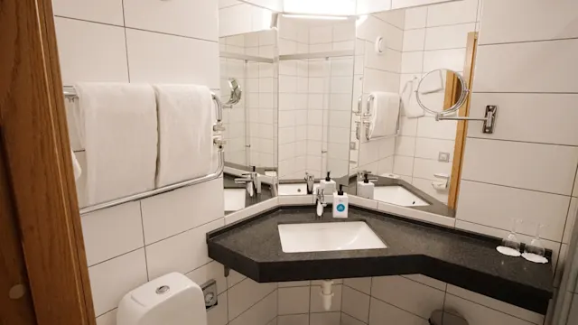 Vitt kaklat badrum, med ett handfat med toalettartiklar, omgiven av flera speglar och handdukshängare. Rummet innehåller en toalett, belyst av stark belysning.