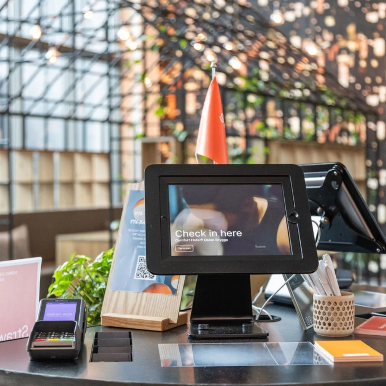 A digital check-in screen displays "Check in here" on a desk with card readers, QR codes, and brochures. A metal structure and a wall with plants are visible in the background.