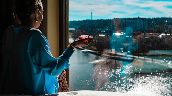 En person i blått håller ett cocktailglas, tittar genom ett fönster på en solbelyst flod och avlägsna byggnader.
