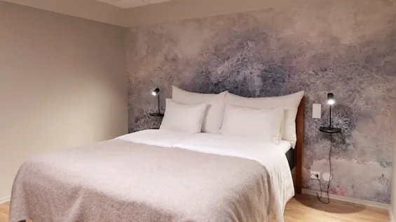 A cozy bedroom featuring a bed with white linens and a light grey blanket, set against a textured, abstract wallpaper.