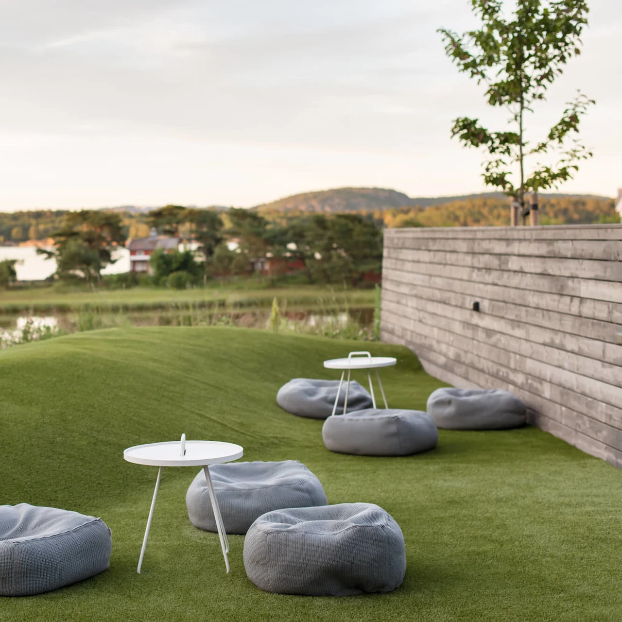 Bean bags and small tables rest on artificial grass hills, bordered by a wooden fence. The backdrop features a serene lake and wooded hills under a cloudy sky.