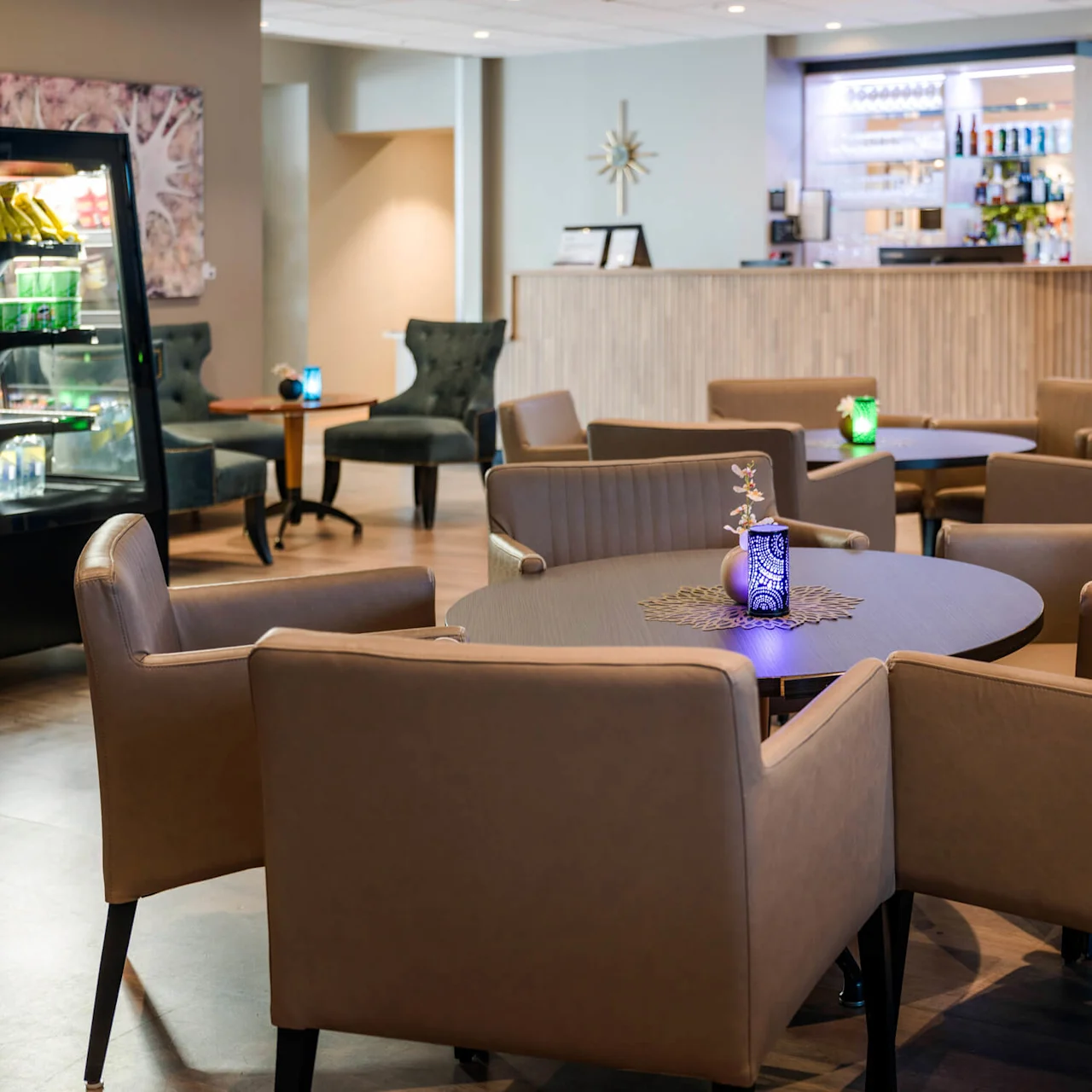 Round table surrounded by beige chairs, set in a modern lounge with a bar in the background. A blue candleholder decorates the table, and a fridge displays snacks nearby.