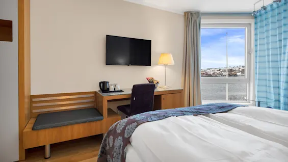 Et koselig hotellrom har en redd seng, en veggmontert TV og et skrivebord med en stol og lampe. Vinduet gir utsikt over vannet og en lys himmel.