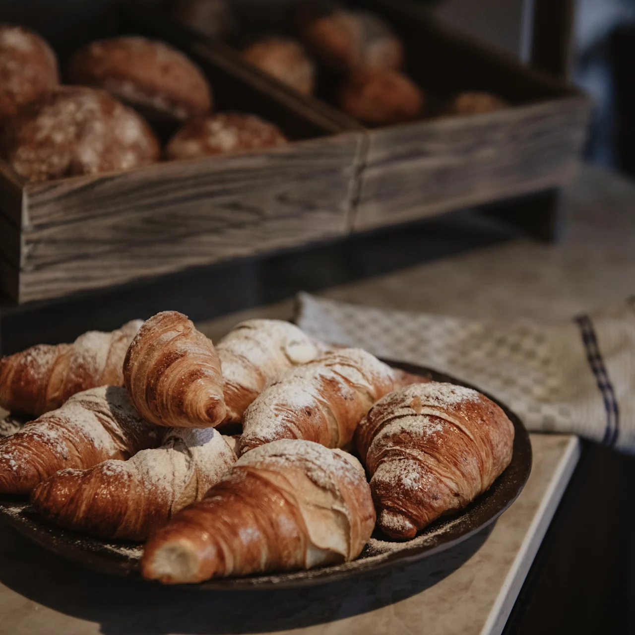 Croissants lie on a round plate, dusted with powdered sugar, on a countertop. Behind them, wooden shelves hold assorted rustic bread rolls, enhancing a cozy bakery setting.