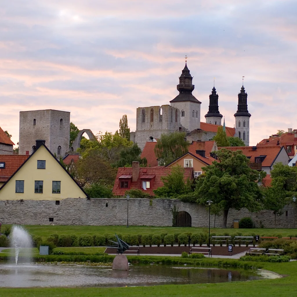A medieval church towers above a town with colorful buildings. A stone wall and leafy trees surround the area. A fountain sprays water in a nearby park pond.