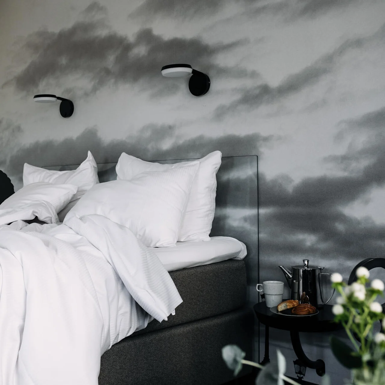 Bed with white linens and pillows is unmade, suggesting recent use. Nearby, a nightstand holds a teapot, cup, croissant, and flowers. Mural of clouds adorns the wall.