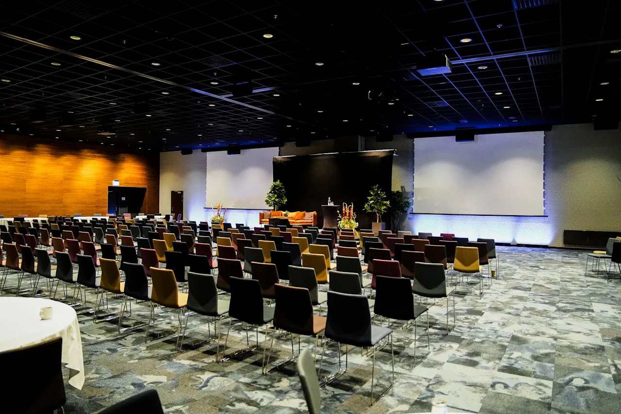 Rader av färgglada stolar är vända mot en stor skärm i ett svagt belyst konferensrum. Scenen är utsmyckad med växter och podier, vilket skapar en formell presentationsmiljö.