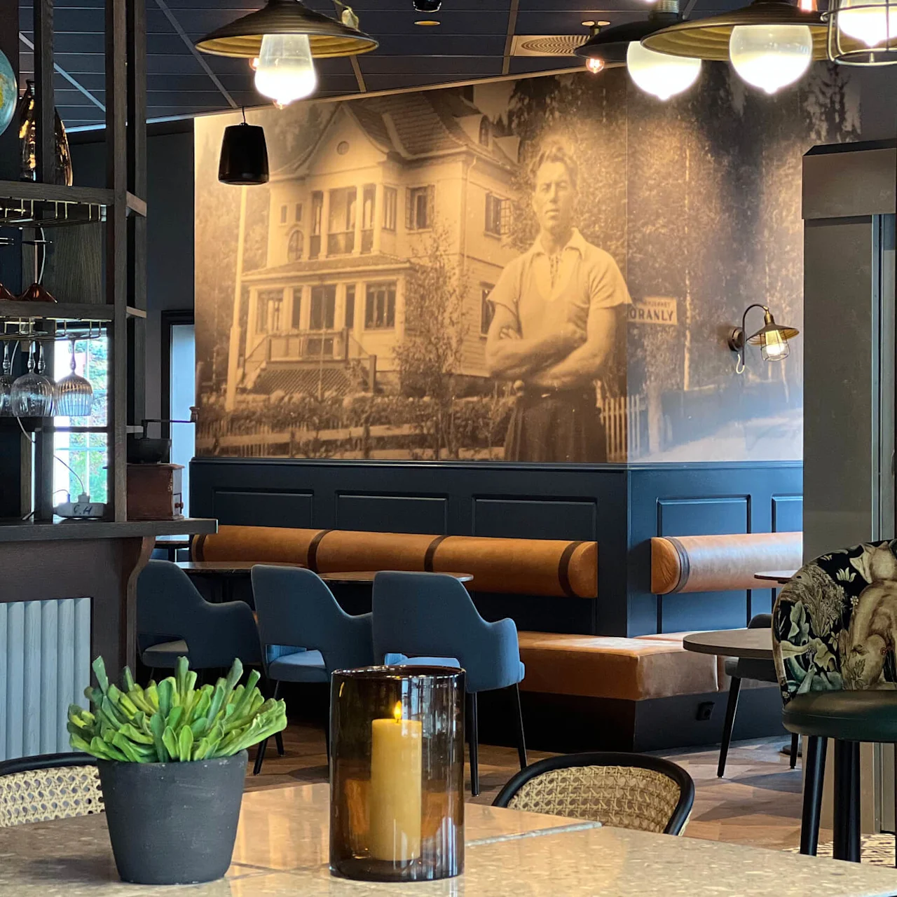 En væg viser et stort vintagefotografi af en mand foran et hus. Caféinteriøret inkluderer hængende lamper, en globus og træmøbler.