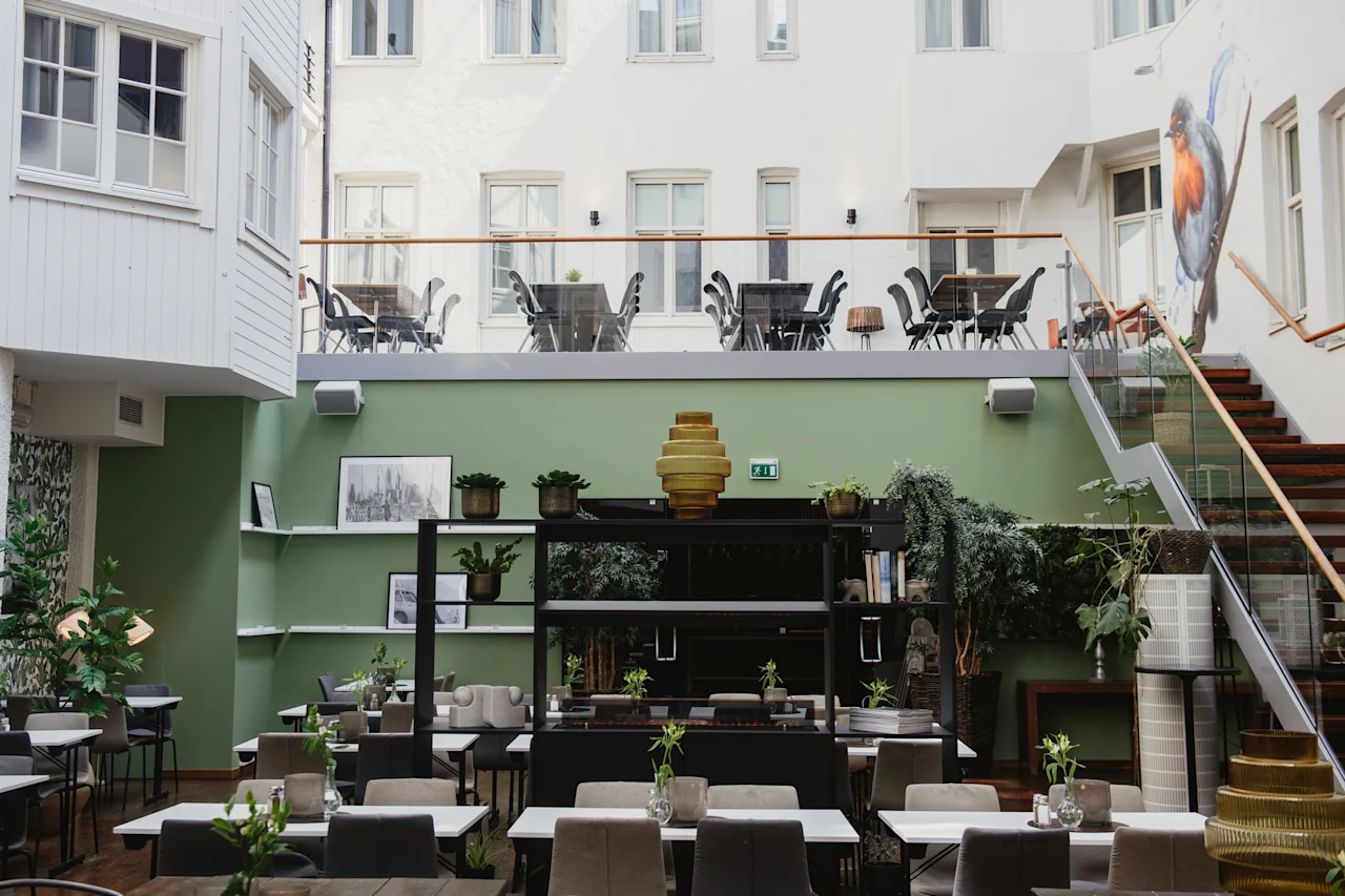 Restaurangbord med växter; omgivande grön inredning och konstverk skapar en modern, luftig atmosfär. Utomhusplatser på övervåningen syns genom en glasbarriär.