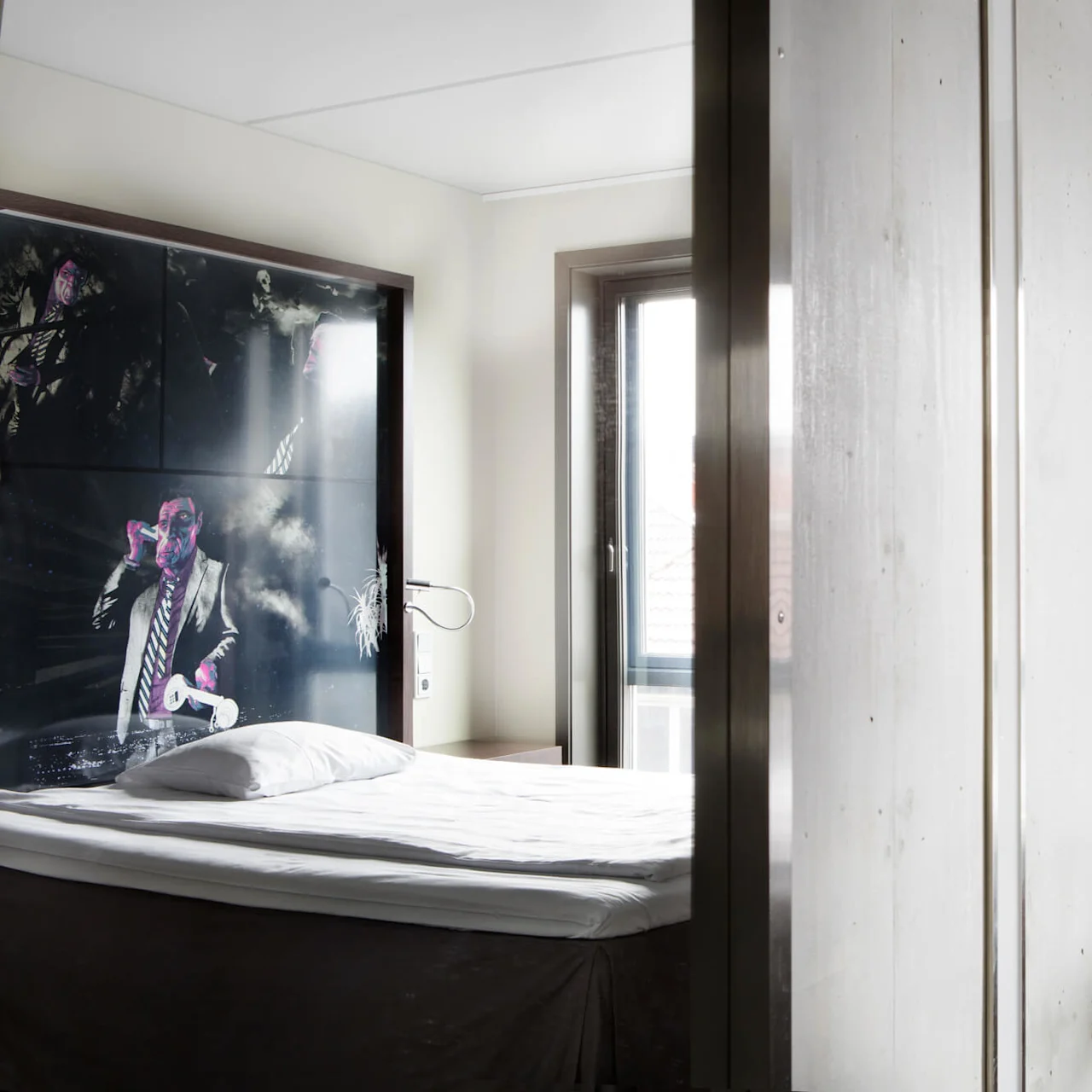 Seng med pute ved siden av kunstverk som viser mann, moderne hotellrom. Glasskrivebord med TV montert på vegg; rød stol; stort vindu med gardin i nærheten.