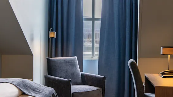 A chair sits beside a window with blue curtains drawn, in a hotel room. A desk with a lamp and phone is nearby. Bed partially visible.
