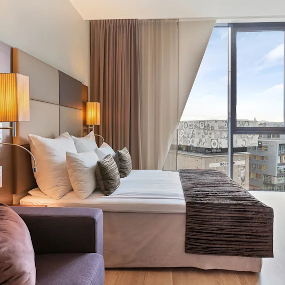 En seng med dekorative puter flankert av veggmonterte lamper i et moderne hotellrom, med utsikt over byens bygninger gjennom et stort vindu.