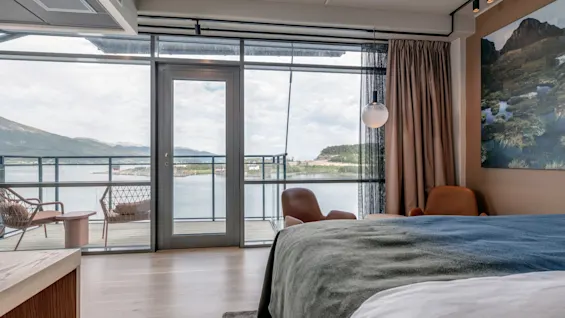 Et hyggeligt hotelværelse med en pænt redt seng og to brune stole, mod store vinduer med udsigt til en altan og en rolig sø omgivet af bjerge.