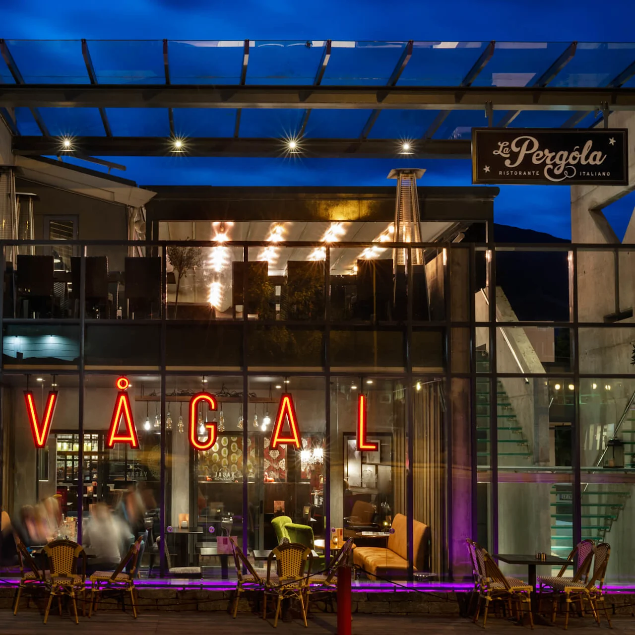 En modern glasfrontad restaurang med röda neonskyltar som stavar "VÅGAL," varmt upplyst inuti, med uteservering. En skylt lyder "La Pergola Ristorante Italiano" under en nattlig himmel.