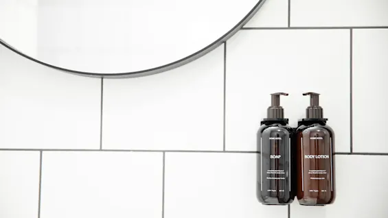 To brune pumpeflasker mærket "SOAP" og "BODY LOTION" monteret på en hvid flisebelagt badeværelsesvæg under et cirkulært spejl.