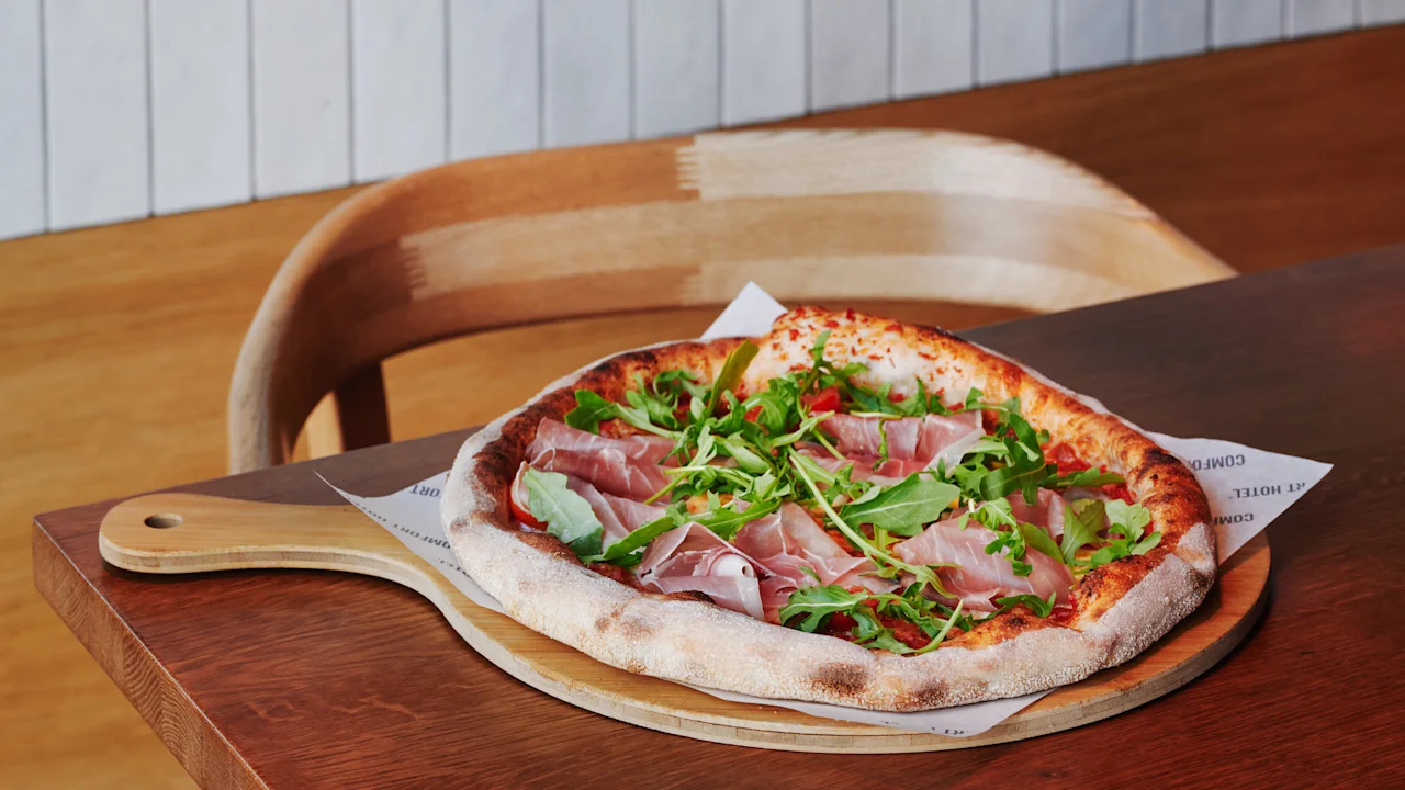Herkullinen pizza ilmakuivatulla kinkulla, rucolalla ja tomaateilla puulapiolla, tarjoiltuna pöydällä. Teksti COMFORT HOTEL.