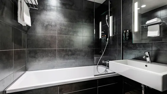 Et badekar med dusjhode står i et stilig, moderne bad. Mørke fliser dekker veggene, og en hvit vask er montert til høyre, under et speil.