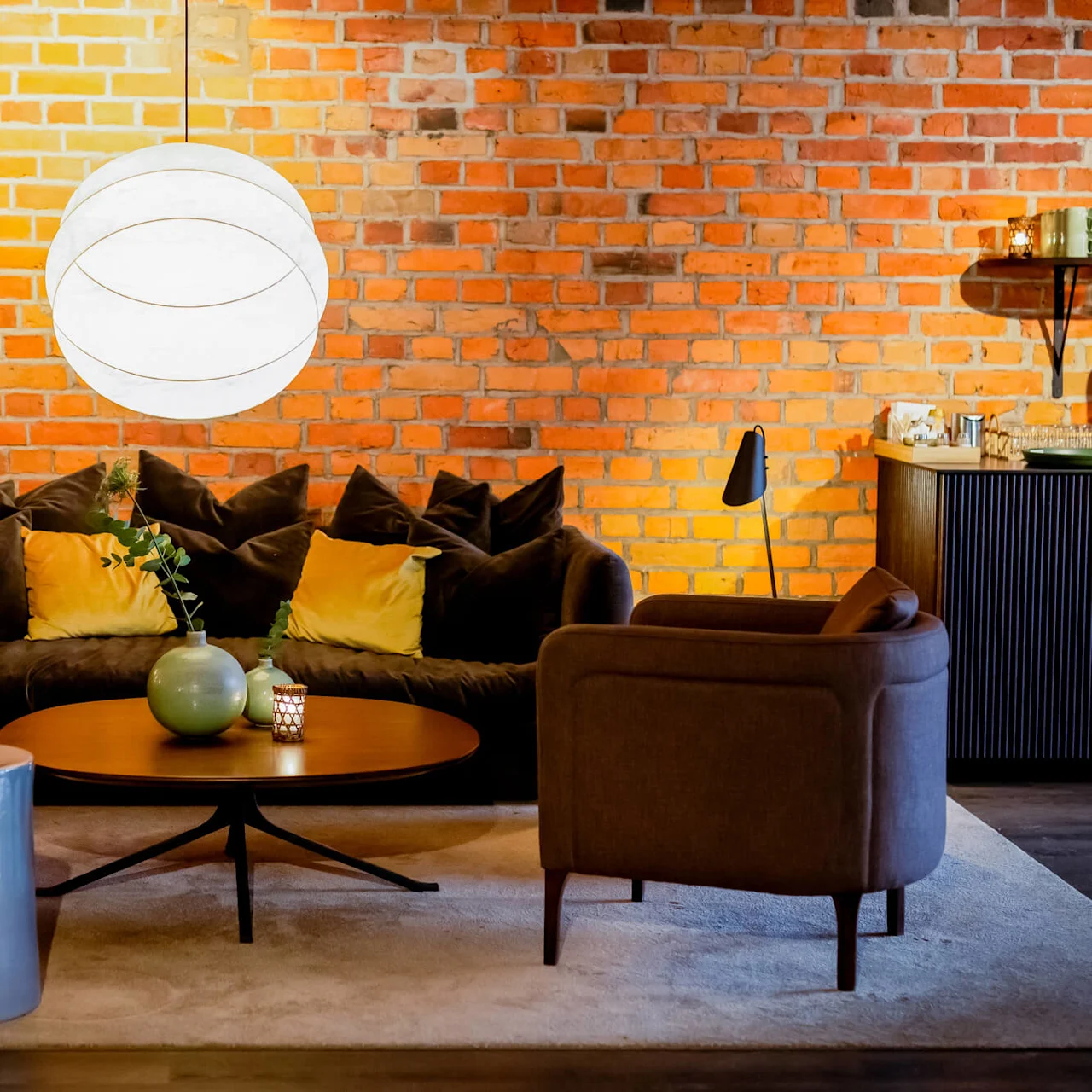 A cozy lounge area with a sofa, armchairs, and a coffee table set against an exposed brick wall, lit by a stylish pendant light.