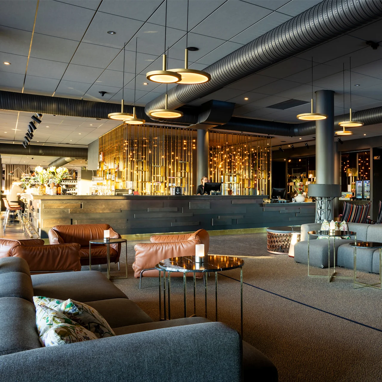 Ett modernt loungeområde har bruna läder- och grå tygmöbler runt låga bord; en elegant bar med gyllene accenter syns i bakgrunden. Färgglad abstrakt konst lyder "we the people."
