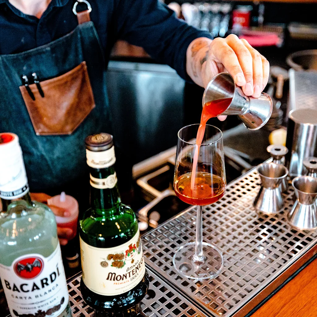 A bartender in an apron pours a red drink from a jigger into a glass on a bar counter with bottles and tools.