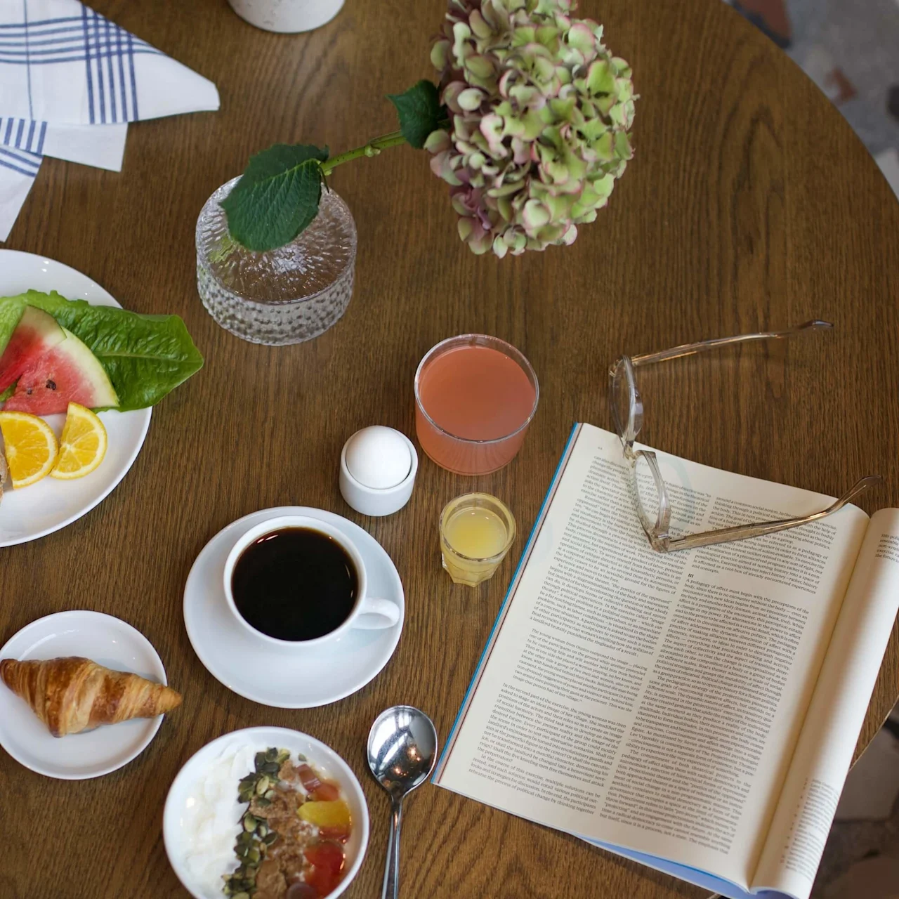 En frukostuppsättning på ett träbord inkluderar kaffe, frukt, yoghurt, en croissant, juice och en öppnad tidskrift med glasögon bredvid. En vas med en rosa blomma tillför dekoration.