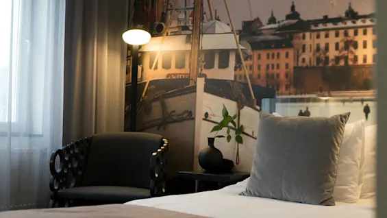 Bed with gray pillow beside modern chair, next to wall mural of a boat and buildings, creating a cozy room ambiance. A soft lamp sheds light on the scene.
