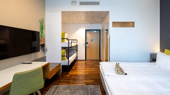 A modern hotel room with a large bed, a desk area, and bunk beds in the background, offering comfortable accommodation.