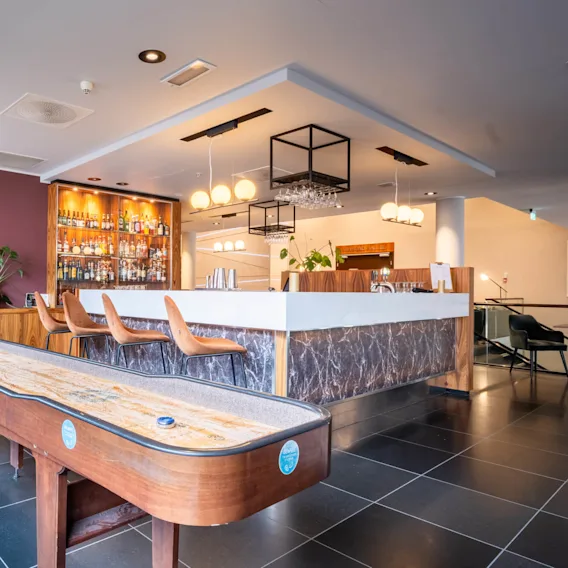 Bar med et shuffleboardbord foran, barstoler og en spritdisplay i en moderne lounge. Koselig belysning skaper en varm atmosfære.