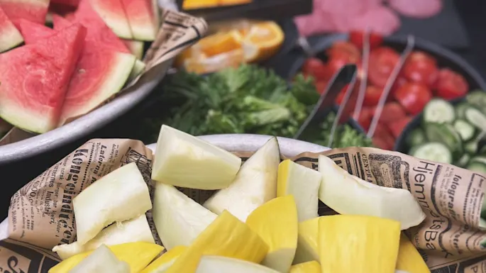 Watermelon and mixed vegetables lie in bowls on a table, surrounded by sliced cucumbers, tomatoes, and greens. Background has more sliced fruits and meats. Newspaper lining is visible.