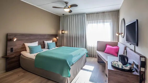 A hotel room with a neatly made double bed, adorned with turquoise accents, is well-lit by natural light from a window, surrounded by modern furnishings and a TV on the wall.