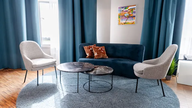 Blue sofa with two pillows, accompanied by two gray chairs and round coffee tables, rests on a circular rug in a room with blue curtains and a wall painting.