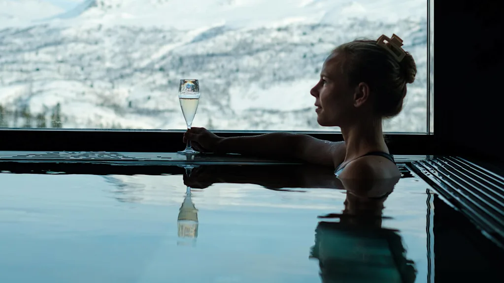En person slapper af i en pool, holder et champagneglas, med sneklædte bjerge synlige gennem et stort vindue.