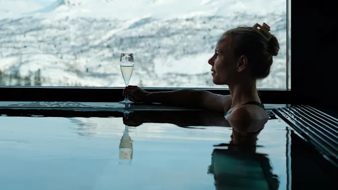 A person relaxes in a pool, holding a champagne glass, with snow-covered mountains visible through a large window.