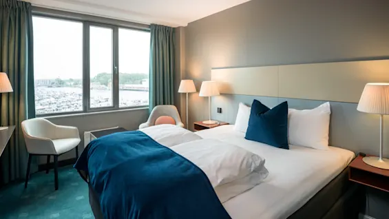 Et moderne hotelværelse med en sirligt redt seng og blå detaljer er oplyst af to sengelamper. Stort vindue afslører en marina med både og grønt.