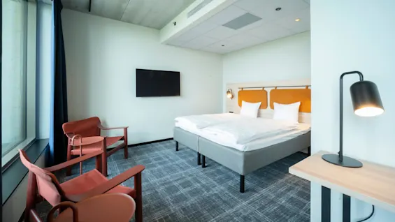 Siististi pedattu parivuode valkoisilla liinavaatteilla sijaitsee modernissa, minimalistisessa hotellihuoneessa. Kaksi punaista tuolia ovat vastapäätä seinälle kiinnitettyä televisiota, valaistuna lattiavalaisimella.