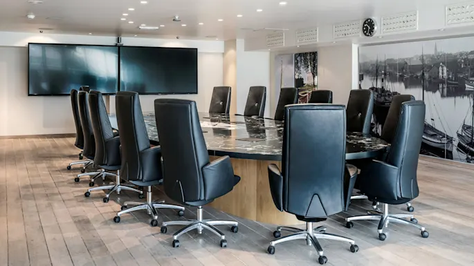 A long, oval conference table is surrounded by black leather chairs on wheels. In the background, there are large screens and murals of a harbor scene.