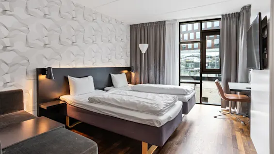 A modern bedroom with two separate beds, textured wallpaper, floor-to-ceiling window, and wooden floor in an urban setting.