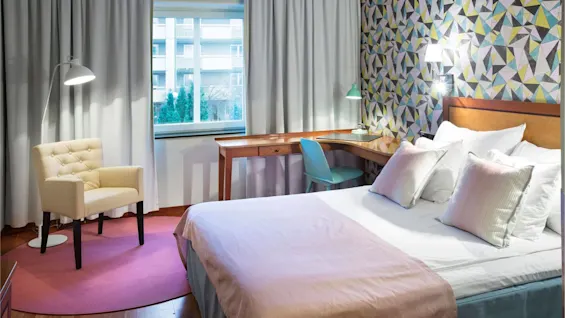 En säng med kuddar vilar i ett rum. En mönstrad vägg, ett skrivbord med lampa, en blå stol och en beige fåtölj på en rosa matta kompletterar den mysiga omgivningen.