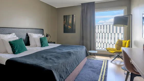 A neatly made bed with green pillows sits in a modern hotel room. A yellow chair and large window provide a view of patterned buildings outside.