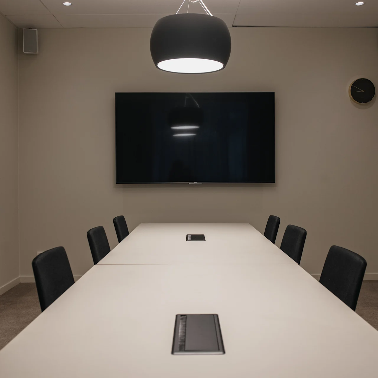 A conference room features a long, white table with black chairs, positioned under a central hanging lamp. A flat-screen TV is mounted on the wall, accompanied by a clock and speakers.