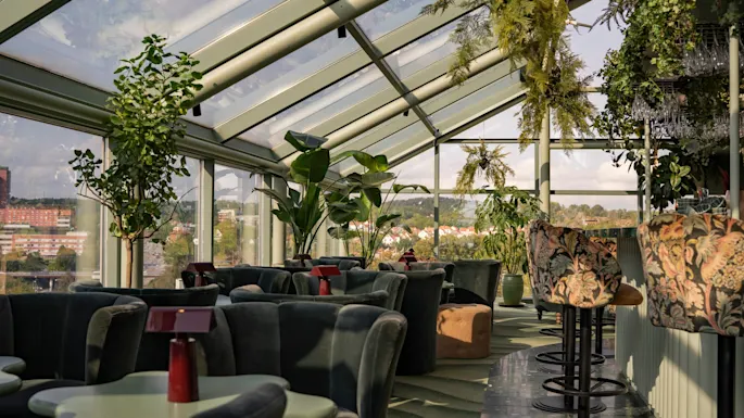 En stilig taklounge med glasstak, frodig grønt og komfortable sitteplasser, med panoramautsikt over byen.