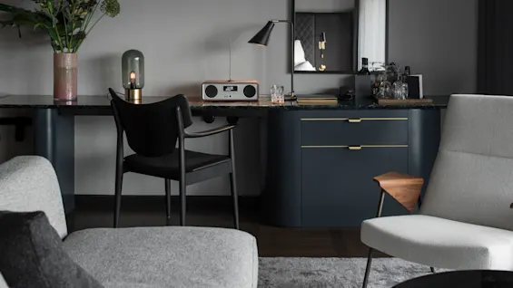 Et skrivebord har en radio, lampe og en vase med blomster nær en stol, omgitt av svakt opplyste moderne møbler og et speil som reflekterer en del av rommet.