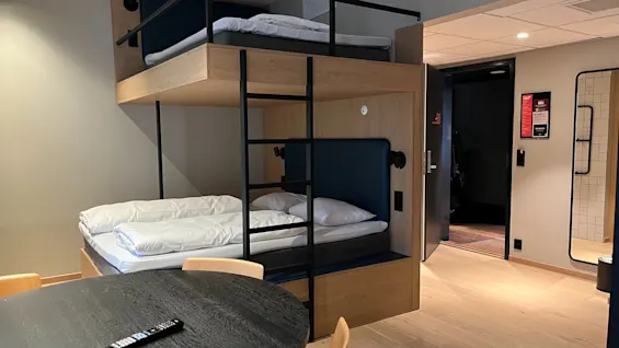 Moderni hotellihuone, jossa kerrossänky, pyöreä pöytä ja avoin ovi. Vaaleat puulattiat ja kyltti, jossa lukee DELI.