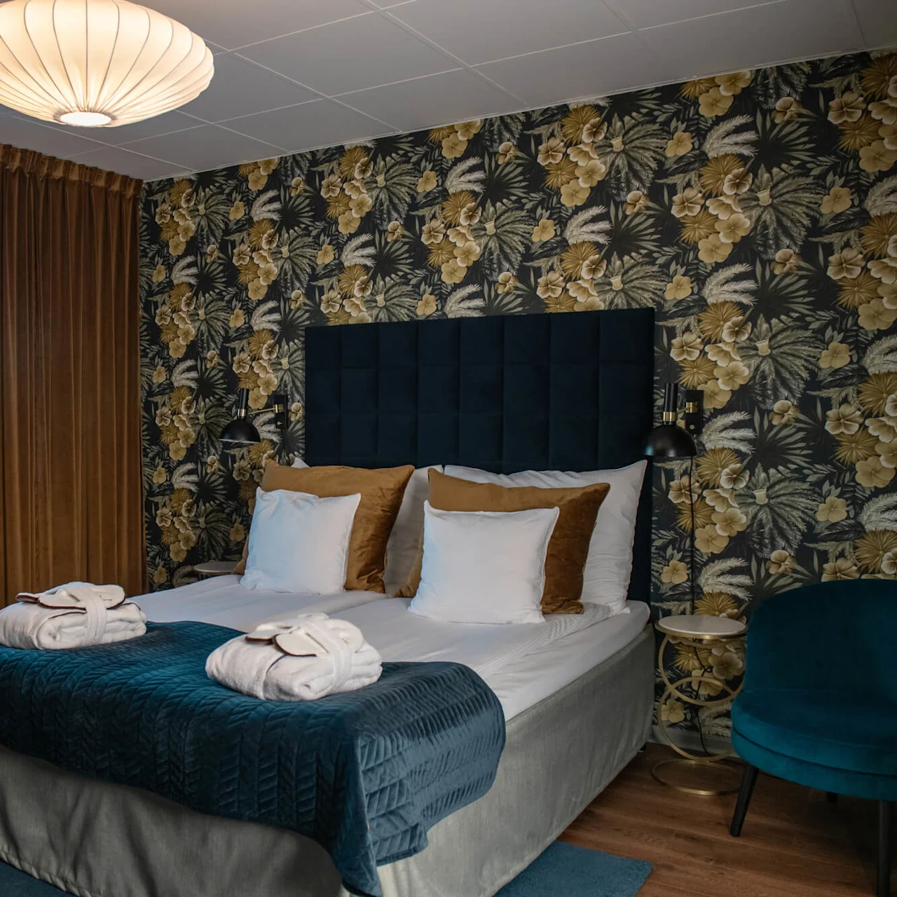 A bed with neatly placed towels rests in a room with floral wallpaper, velvet drapes, and a plush chair, illuminated by a ceiling light.