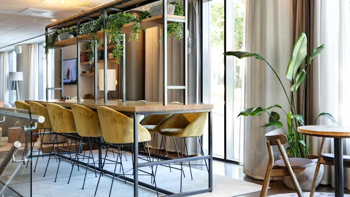 A long wooden table with yellow chairs stands in a bright room, adorned with hanging green plants and shelves, next to tall windows with flowing curtains and a large potted plant.