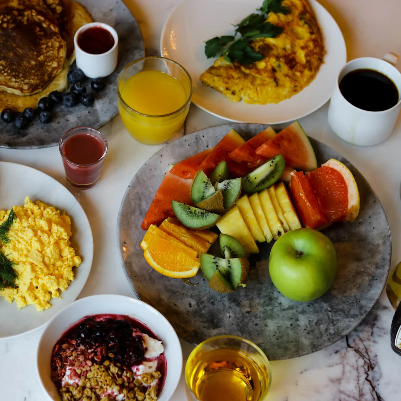 Various breakfast dishes are served on a marble table, including pancakes with blueberries, scrambled eggs with salmon, an omelette with herbs, fresh fruit, assorted breads, and beverages like juice and coffee.